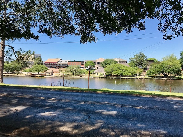 River bank lined with shade trees, a road, and buildings in Natchitoches, Louisiana.
